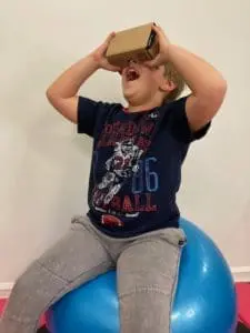 A young boy wearing a dark blue shirt with a football graphic sits on a blue exercise ball, holding a Google Cardboard viewer to his eyes.