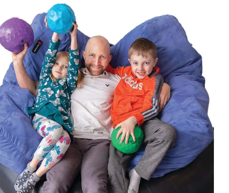 A man sits on a large blue beanbag chair with two children, each holding a colorful ball.