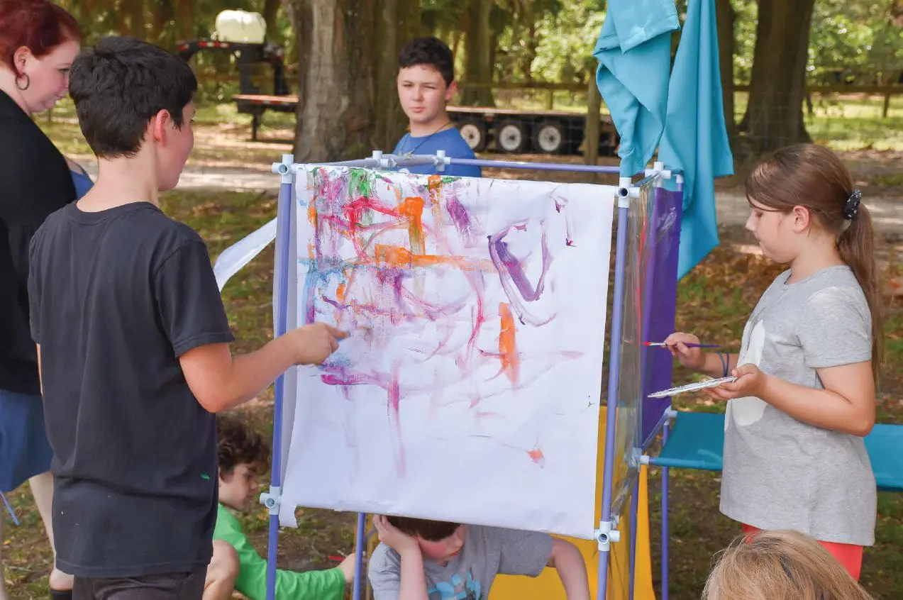 Children paint on a large sheet of paper displayed on an easel outdoors.