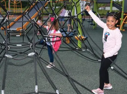 Ava Binion waving while Alivia Thompson climbs on a rope structure at an inclusive playground.