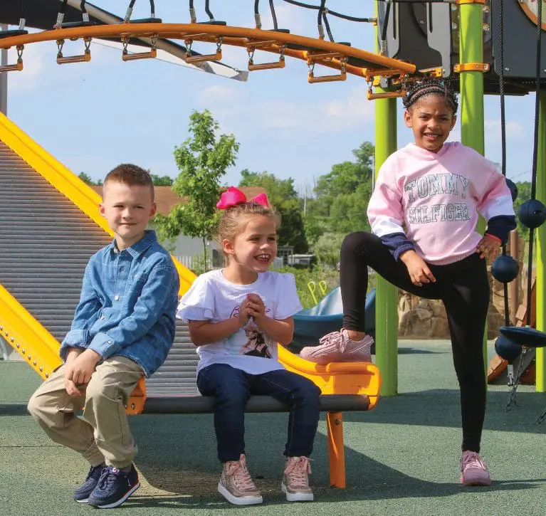 Three children sitting and standing on a playground, with a yellow slide behind them.