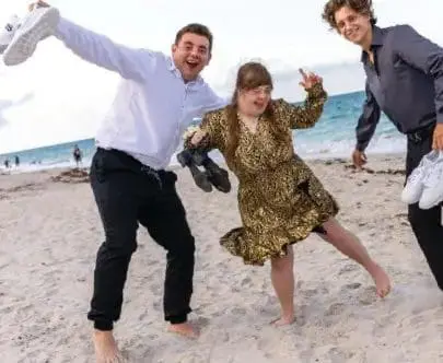 Three young adults pose on a beach, two men and one woman, with the woman in the middle.