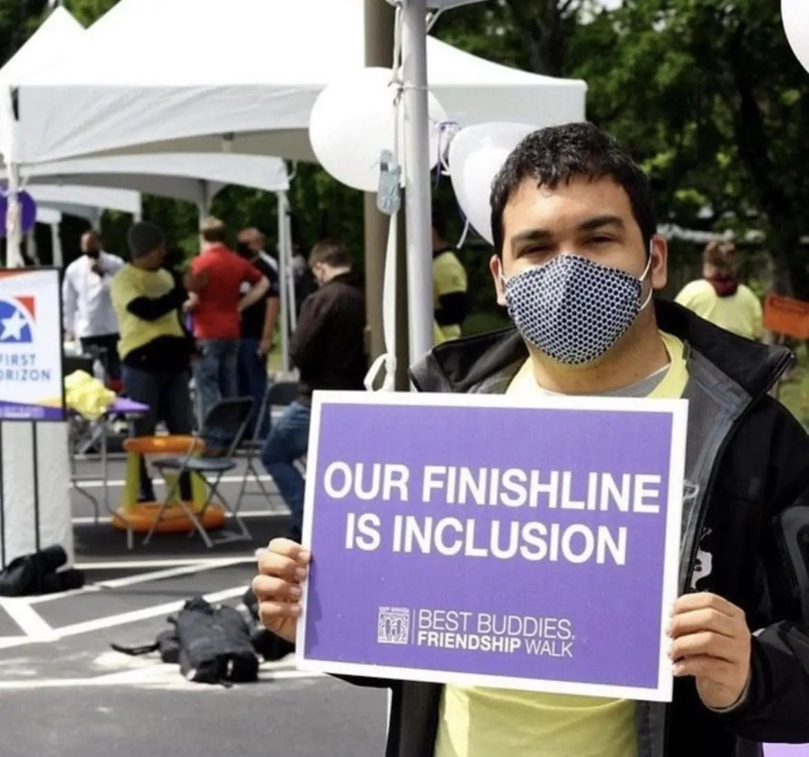 A young man wearing a mask holds a purple sign that says "OUR FINISH LINE IS INCLUSION".