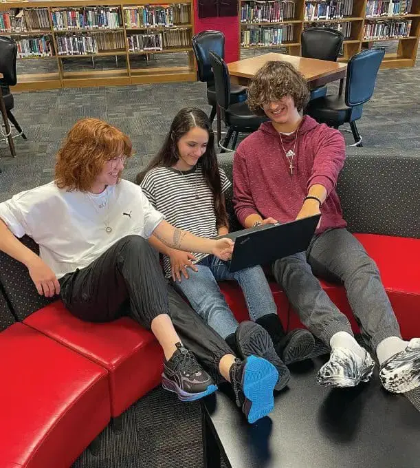 Three students collaborate around a laptop on a red couch in a library.