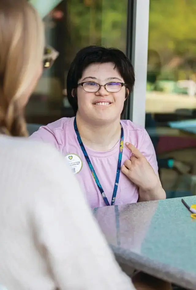 A young woman with glasses and a purple shirt smiles while looking at someone across a table.