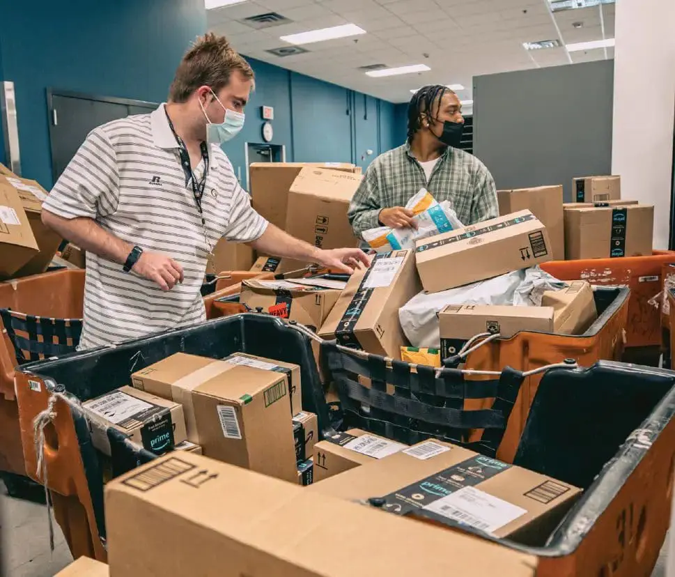 Two men wearing masks sort packages in a shipping facility.