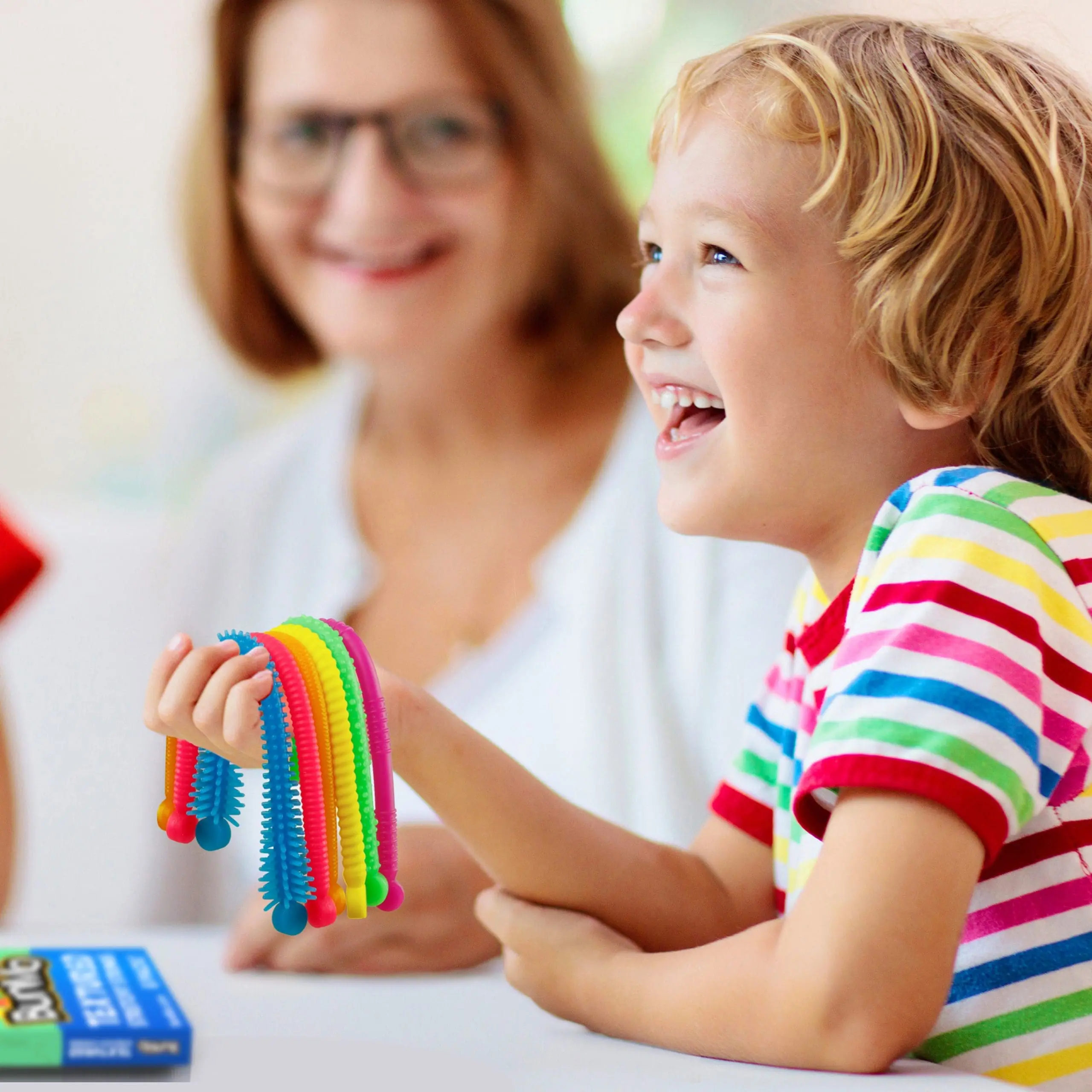 A child smiles while holding a rainbow-colored BunMo fidget toy, with an adult blurred in the background.