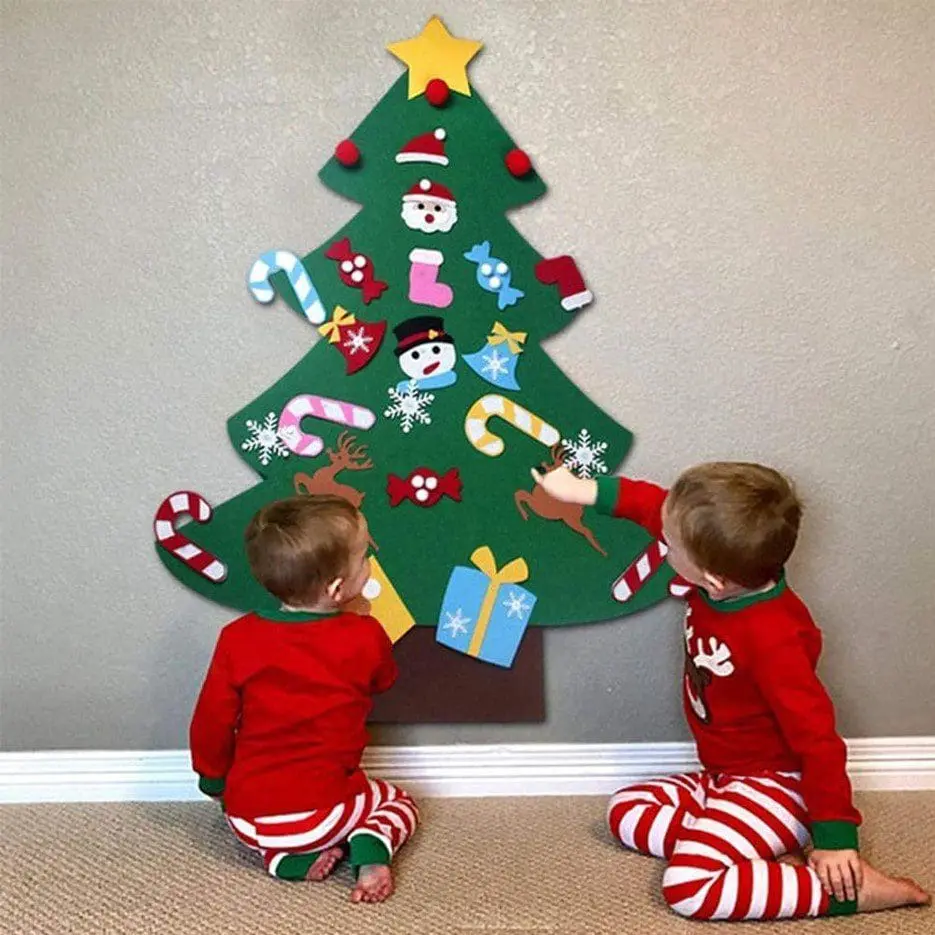 Two children in pajamas decorate a green felt Christmas tree hanging on a wall.
