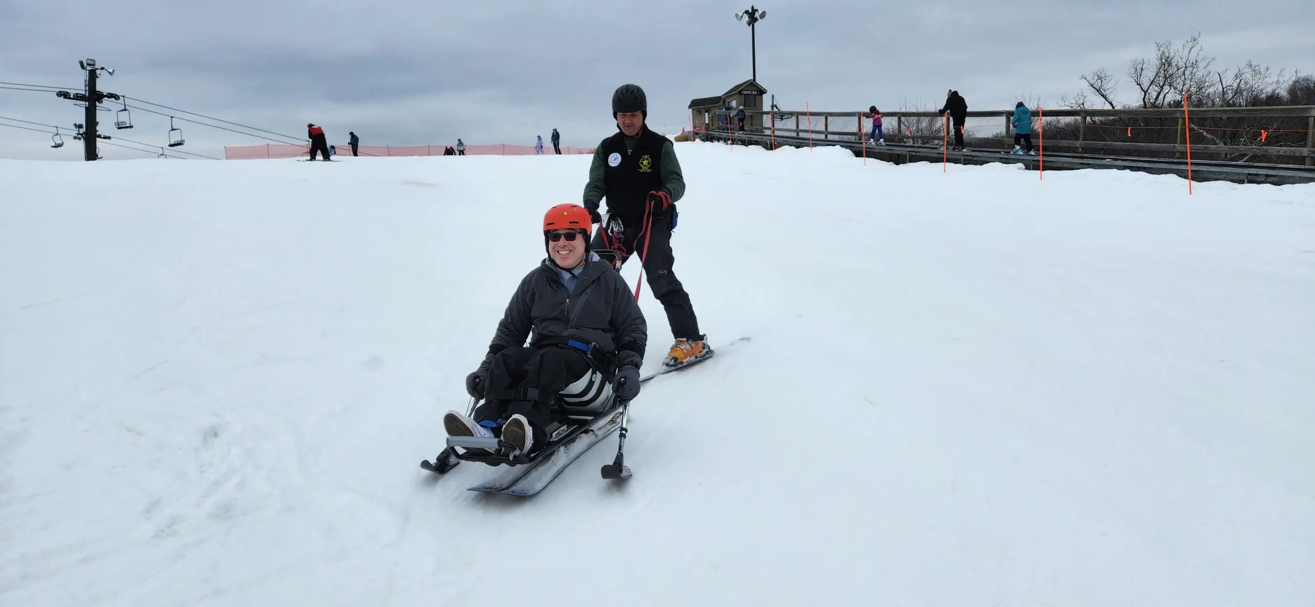 Two people ski down a snowy slope, one on a sit-ski with an instructor behind them.