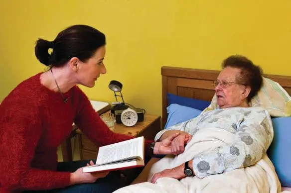 A caregiver reads a book to an elderly woman in bed.