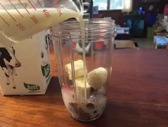 Milk is poured from a measuring cup into a blender with banana chunks.
