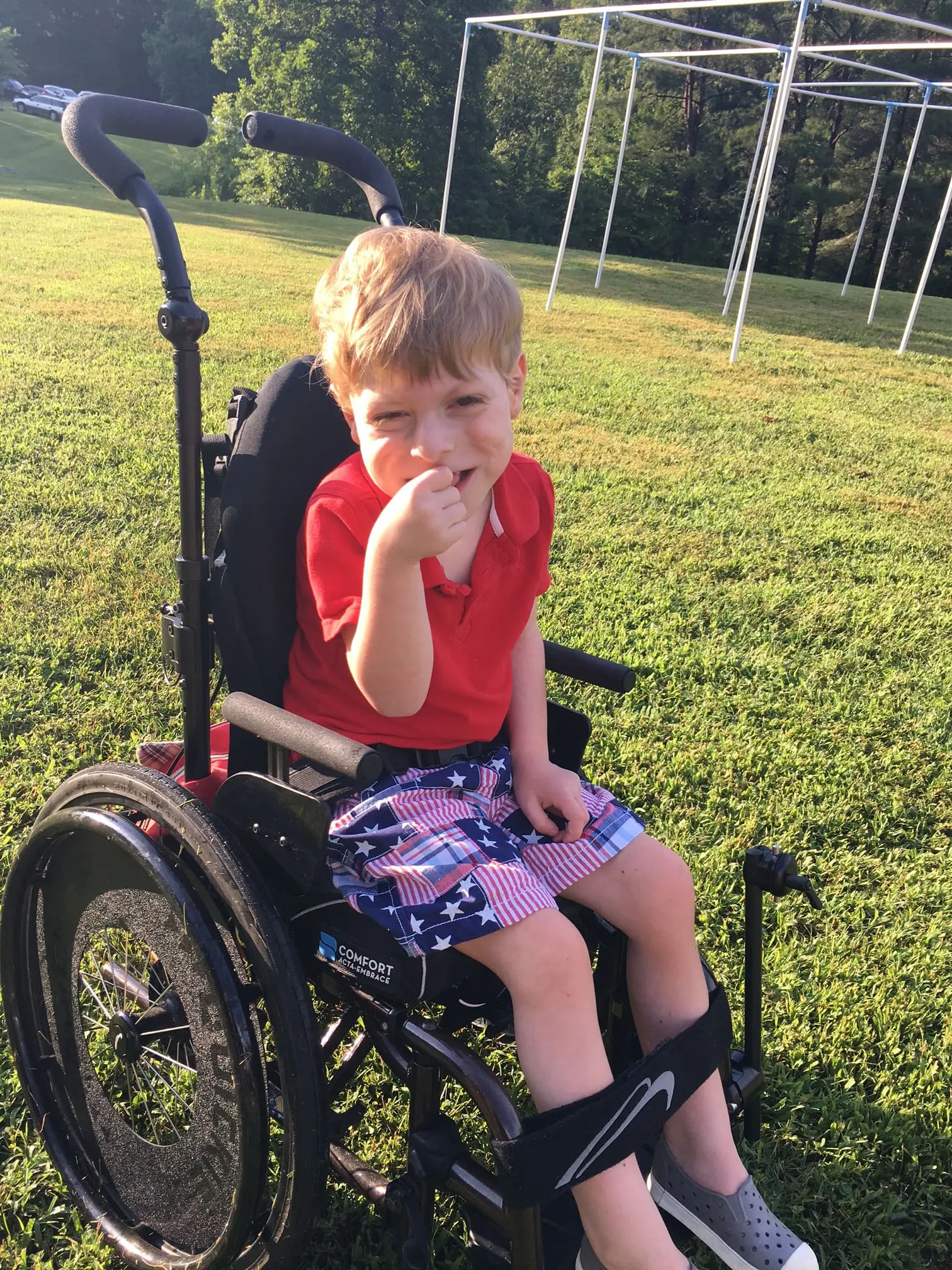 Young boy sitting in a wheelchair outside on a grassy field.