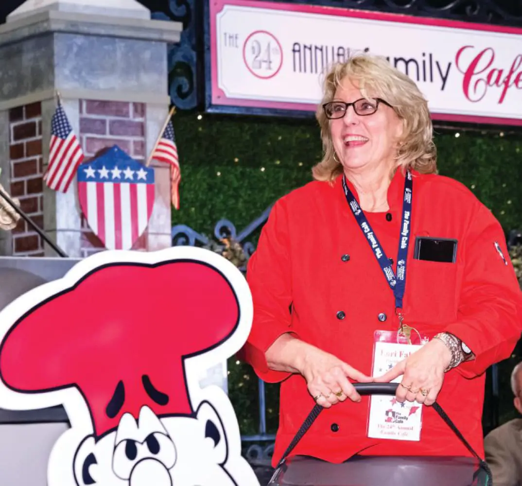 A woman in a red chef's coat smiles while holding a sign with a cartoon chef hat.