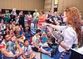 Woman with long curly hair points to a large group of people sitting on the floor.
