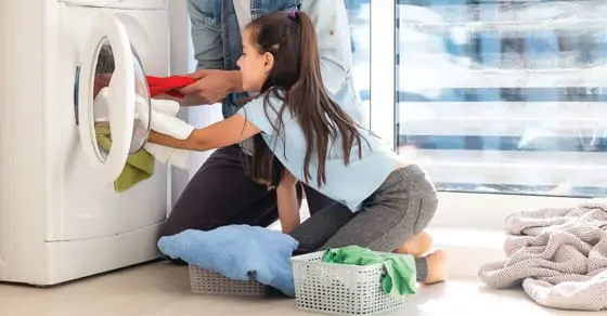 Child helps load laundry into a washing machine next to an adult.