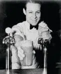 A smiling soda jerk offers a foamy beverage from a soda fountain.