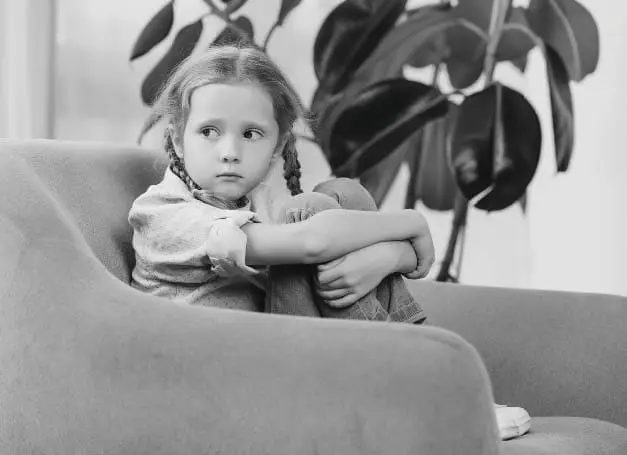 A young girl sits with her arms wrapped around her knees in an armchair.