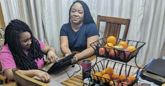 Two women sit at a table with fruit baskets, a tablet, and books.