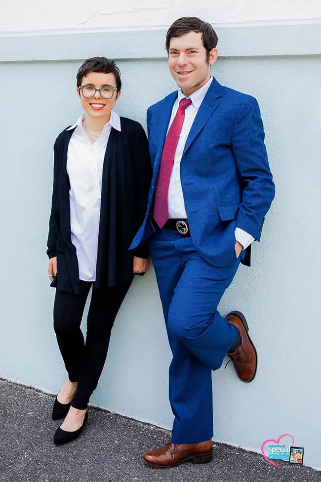Bradley in a navy suit, white shirt, red tie, and brown loafers stands next to a woman in a black cardigan over a white shirt.