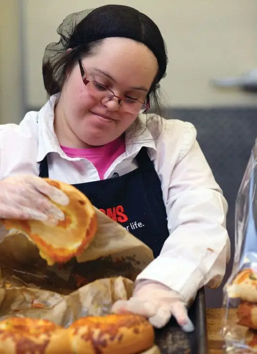 A young woman wearing an apron and hair net prepares baked goods.