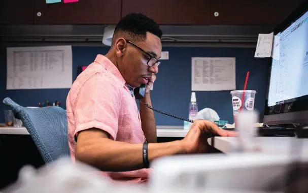 Man in a pink shirt talking on the phone at a desk.