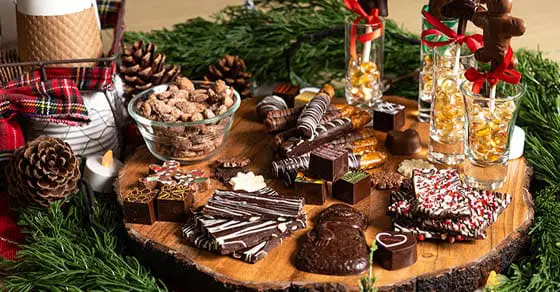 A chocolate charcuterie board with various candies and nuts displayed on a wooden slice surrounded by pine branches.