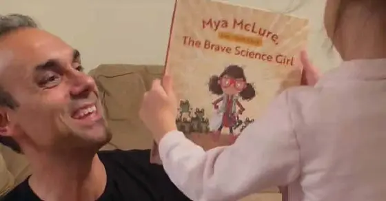 Man smiles as a child holds up a book titled "Mya McLure, The Brave Science Girl.