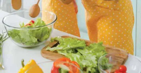 Bowl and wooden cutting board with lettuce and vegetables, with people in yellow polka-dot aprons in the background.