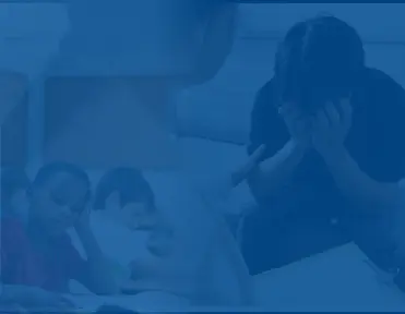 A person sits with their head in their hands while two children appear to be struggling with schoolwork.
