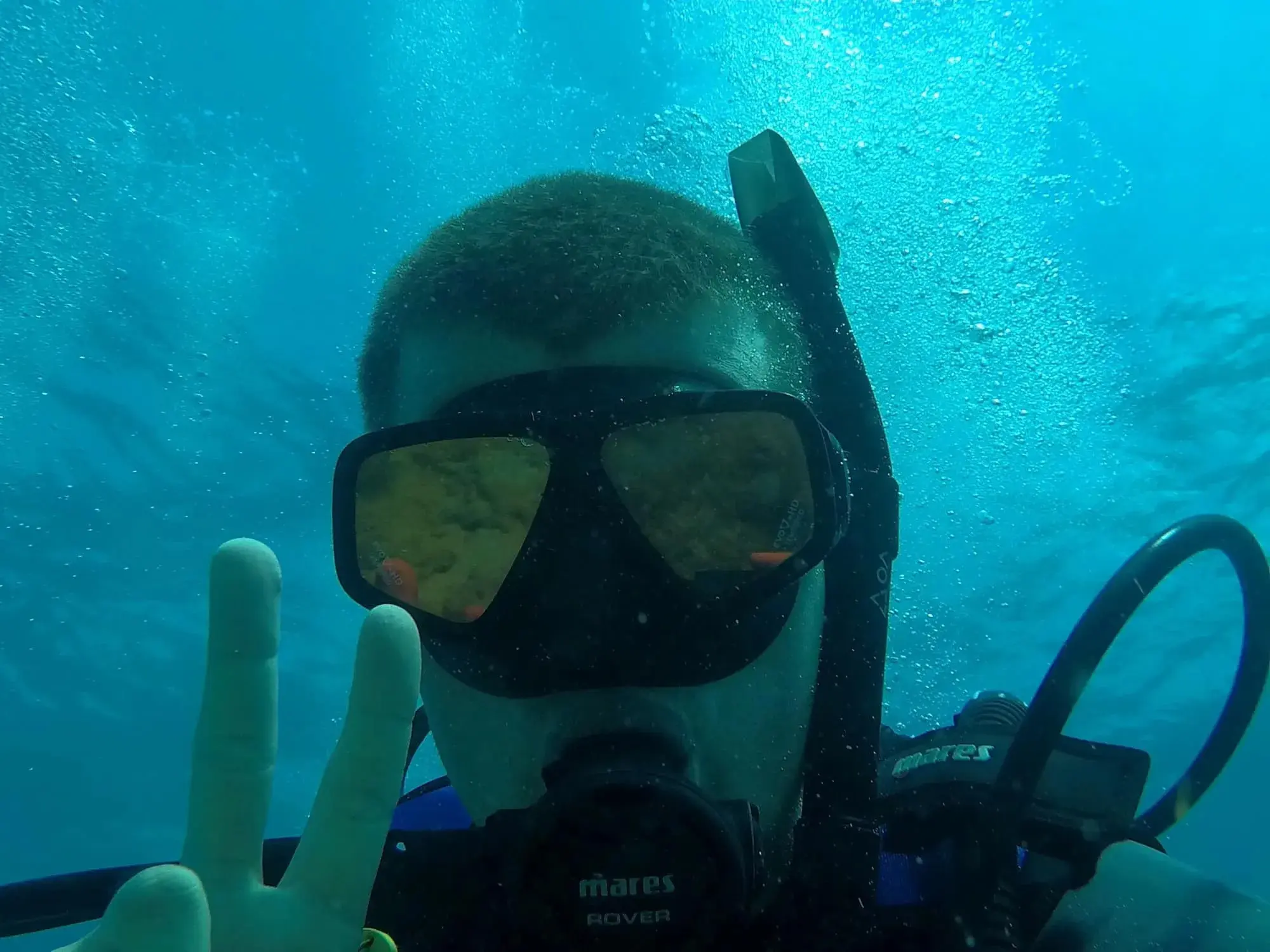 Clayton in a diving mask and scuba gear gives a peace sign underwater.