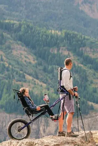 A man with trekking poles stands next to a Huckleberry Hiking device carrying a child on a mountain trail.