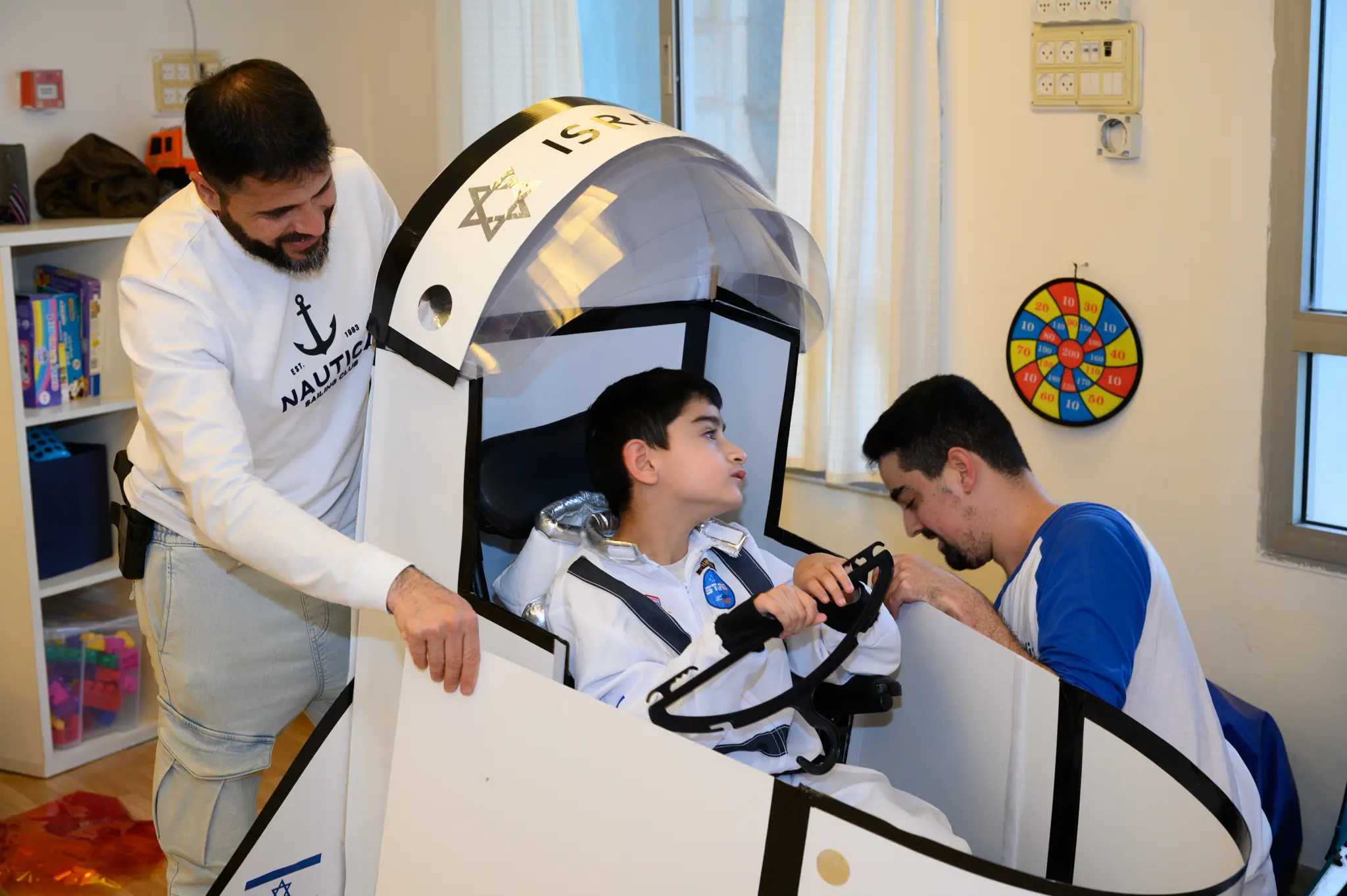 A boy in a wheelchair is dressed as an astronaut inside a rocket ship costume with two men on either side.