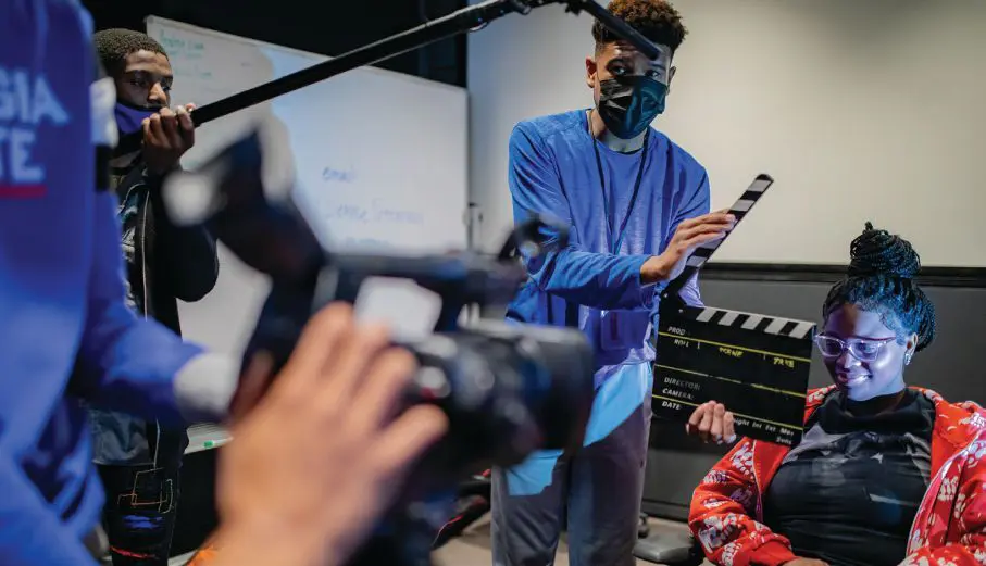 Students work on a film set with a camera, boom mic, and clapperboard.