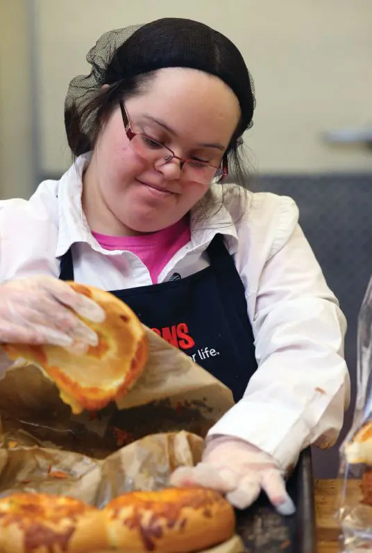 A person wearing an apron and hair net prepares baked goods.