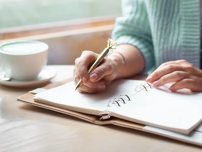 Person writing in a notebook next to a cup of coffee