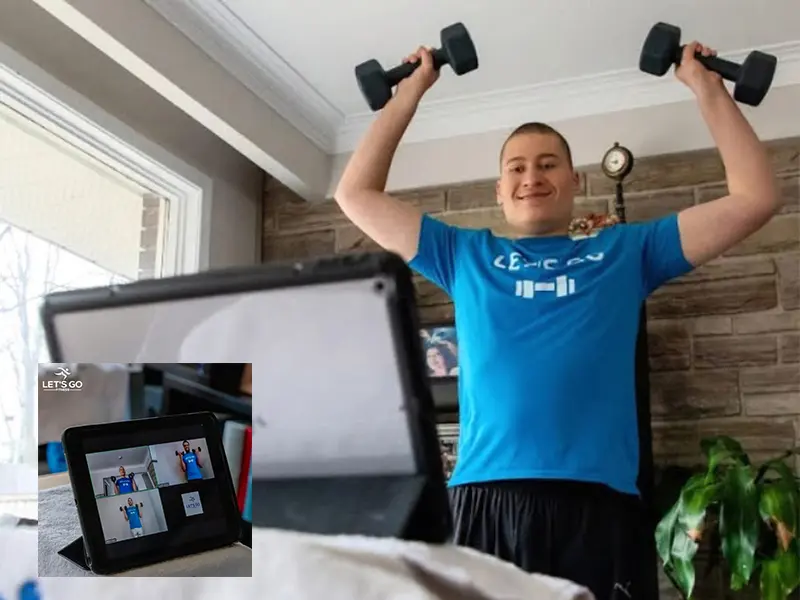 A young man in a blue t-shirt lifts dumbbells while looking at a laptop and tablet.
