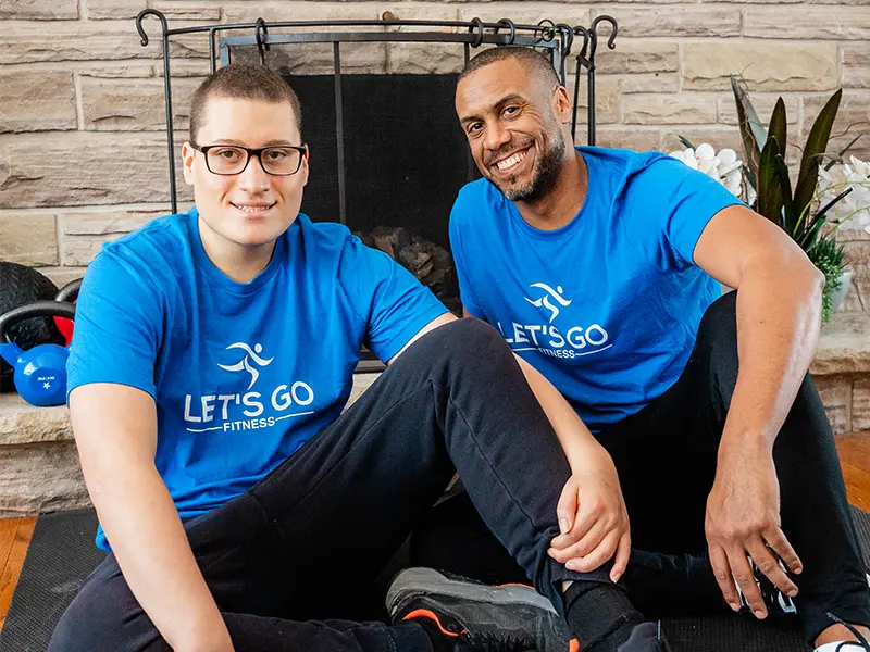 Keegan Johnson and Dante Johnson sitting on the floor in front of a fireplace wearing blue Let's Go Fitness shirts.