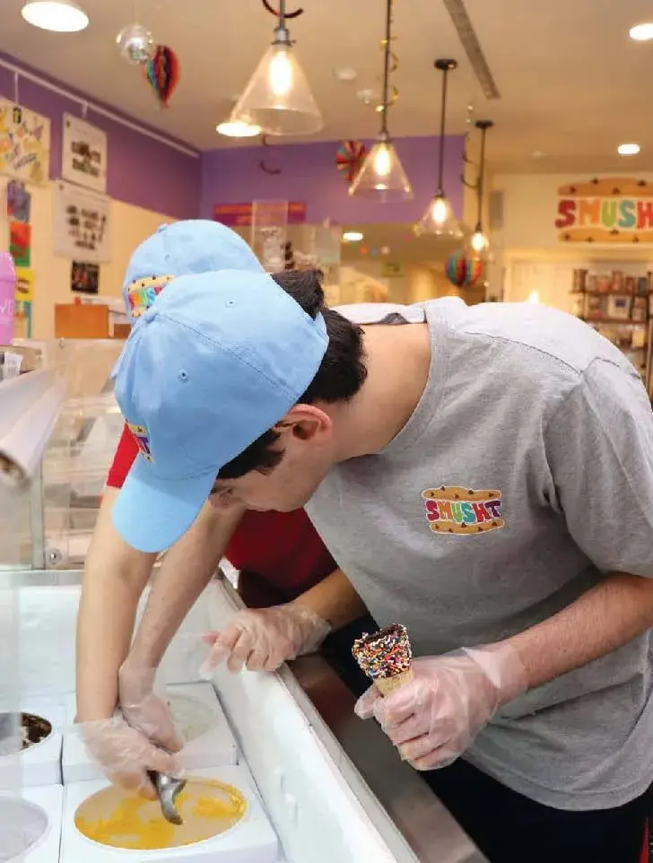Two individuals wearing blue baseball caps and gloves working together to scoop ice cream behind a counter.