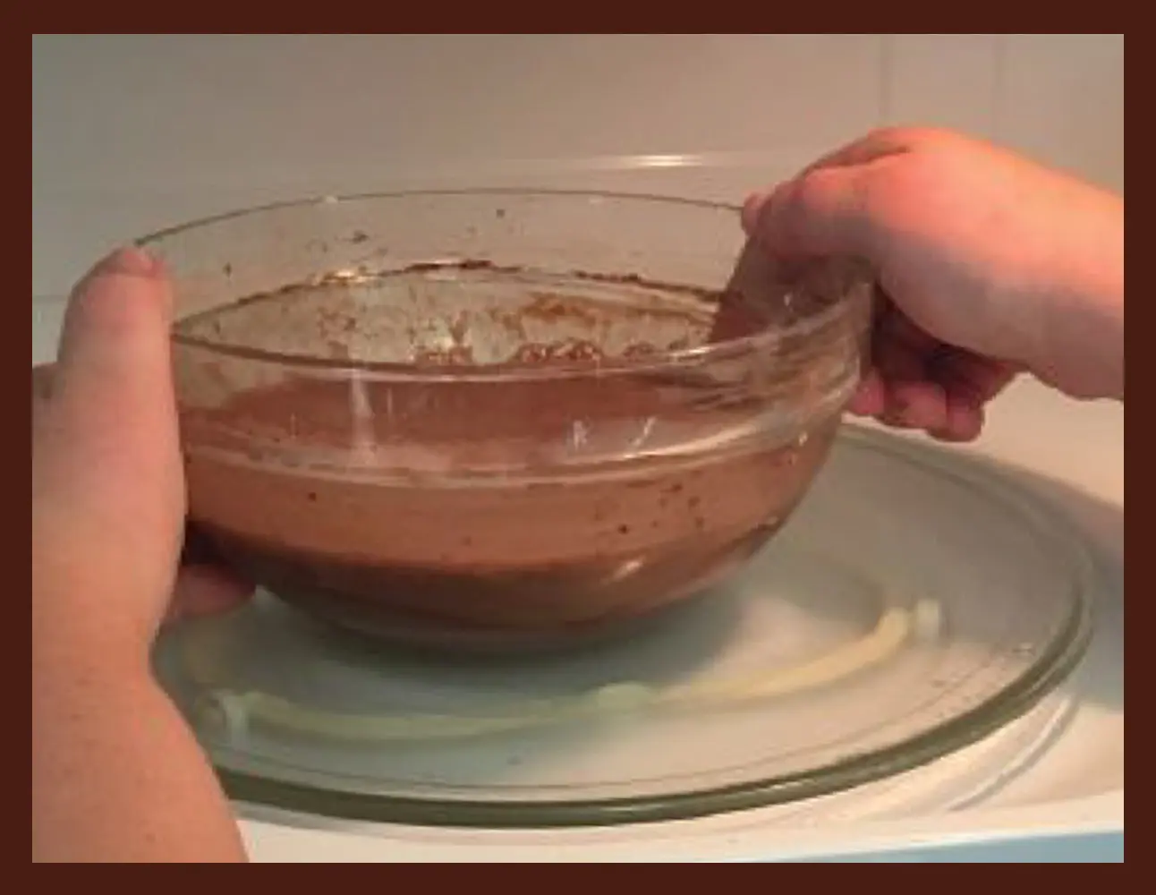 Hands mixing chocolate pudding ingredients in a glass bowl on a plate.