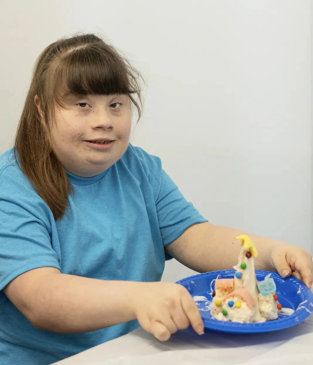 A young person with Down syndrome in a blue shirt decorates a mini edible village on a blue plate.