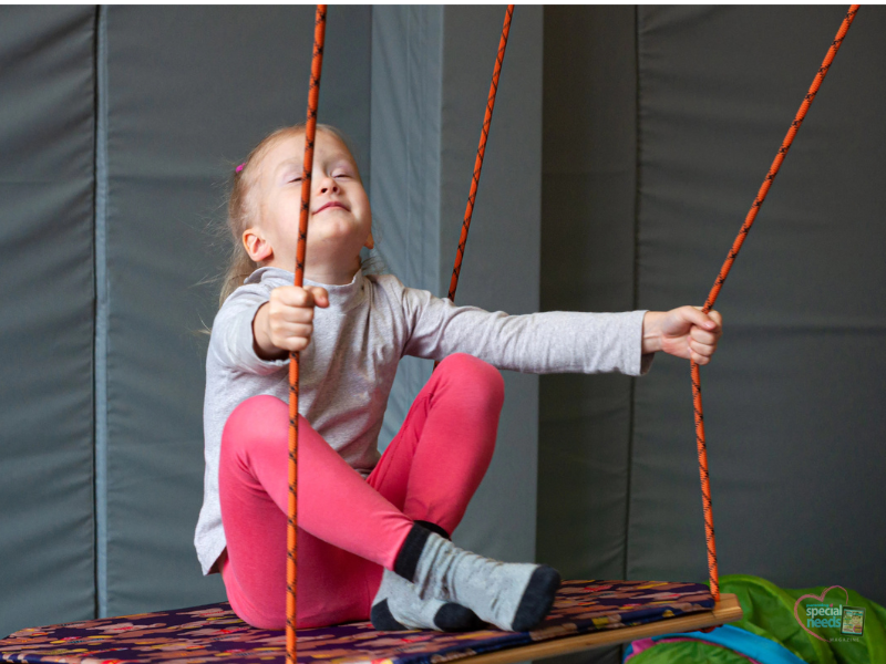 A young girl in a grey long-sleeved shirt and pink leggings sits on a swing, holding the ropes with her eyes closed.