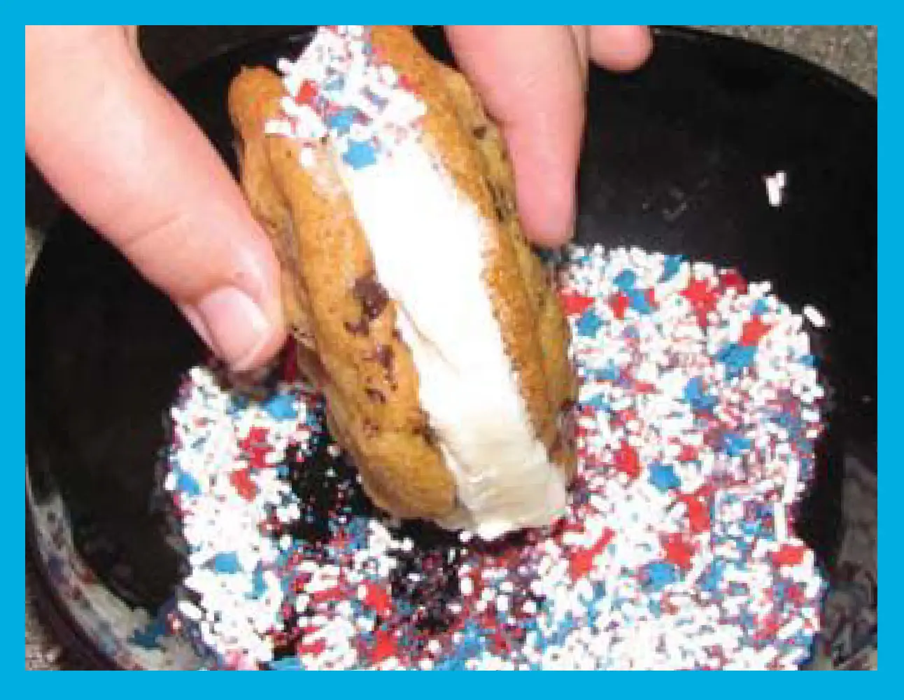 Hand dips an ice cream sandwich into patriotic sprinkles.