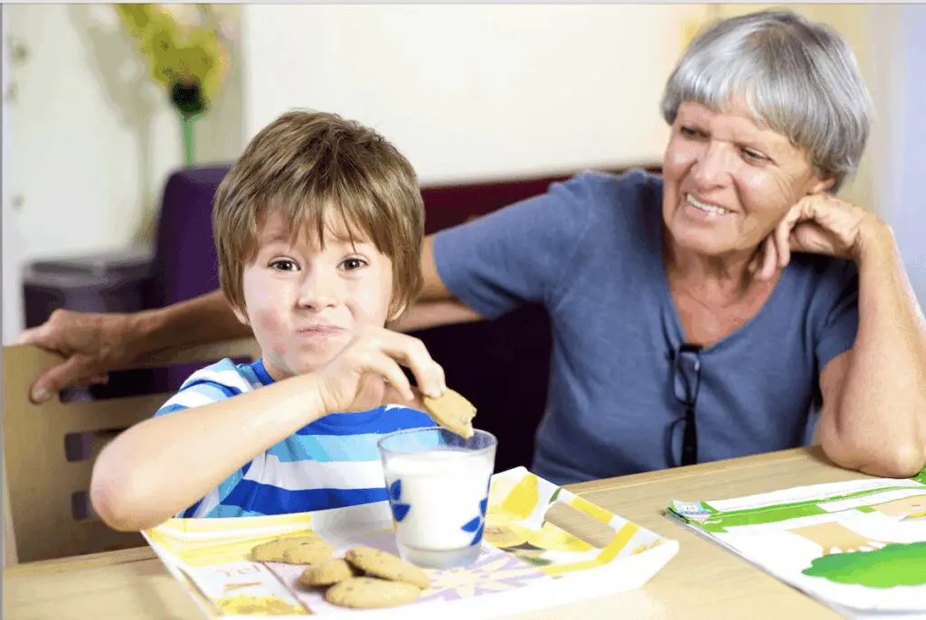 A young boy dips a cookie into milk while sitting with a woman.