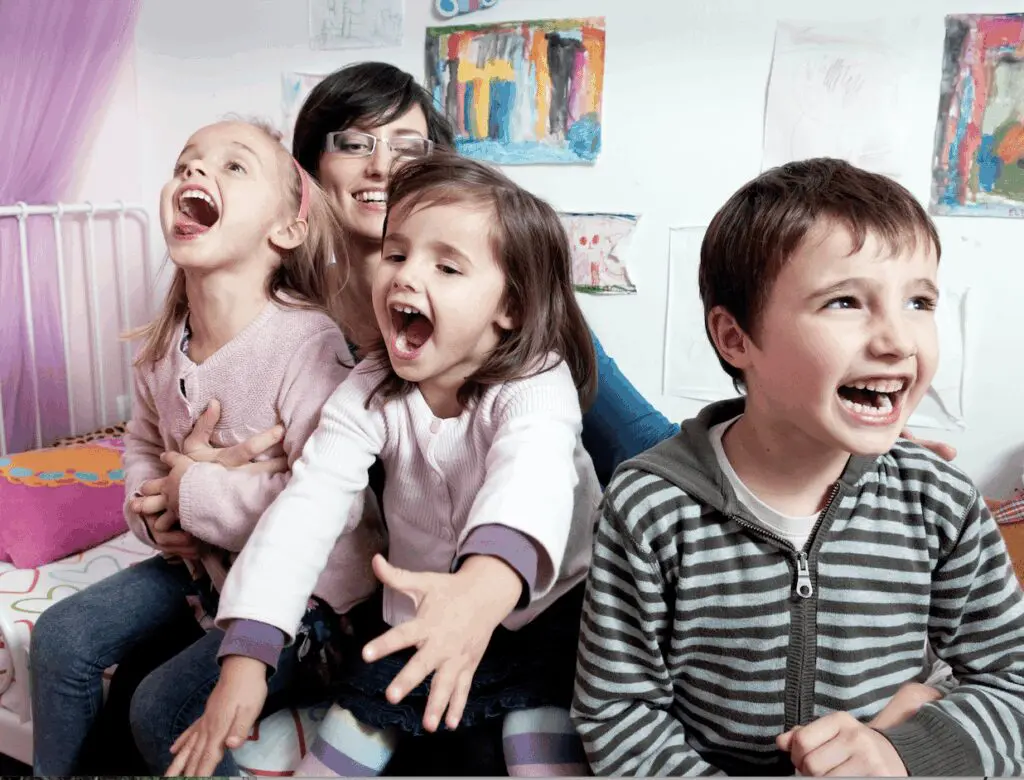 A woman and three children laugh together in a room with children's artwork on the wall.