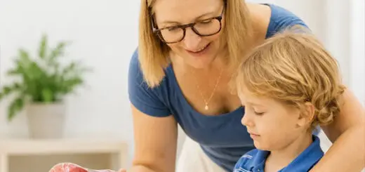 In the image, a woman and a child are engaged in an activity involving a red apple. The woman, dressed in a blue shirt, is holding the apple with both hands, presenting it to the child. The child, wearing a blue shirt as well, has his gaze fixed on the apple, seemingly intrigued by its presence. They are situated indoors, with a white wall serving as their backdrop. A potted plant adds a touch of greenery to the scene. The image does not contain any discernible text. The woman's action of presenting the apple and the child's focused gaze suggest an interaction centered around this object