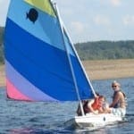 Two people sail on a colorful sailboat on a lake with wooded hills in the background.
