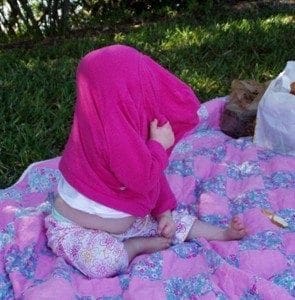 Baby sitting on a blanket with a pink shirt over its head