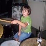 A young boy with curly hair plays a drum set in a room with a fan in the background.
