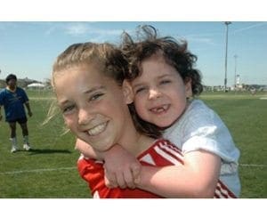 Two girls smile while one carries the other on her back on a soccer field.