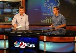 Two men smile at a news desk with a Special Olympics logo in the background.